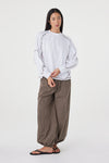 Model wearing the cream long-sleeve tee with brown check trousers and black sandals, showing the dropped shoulders, ribbed cuffs, curved sleeve seams and relaxed drape in a styled front view.