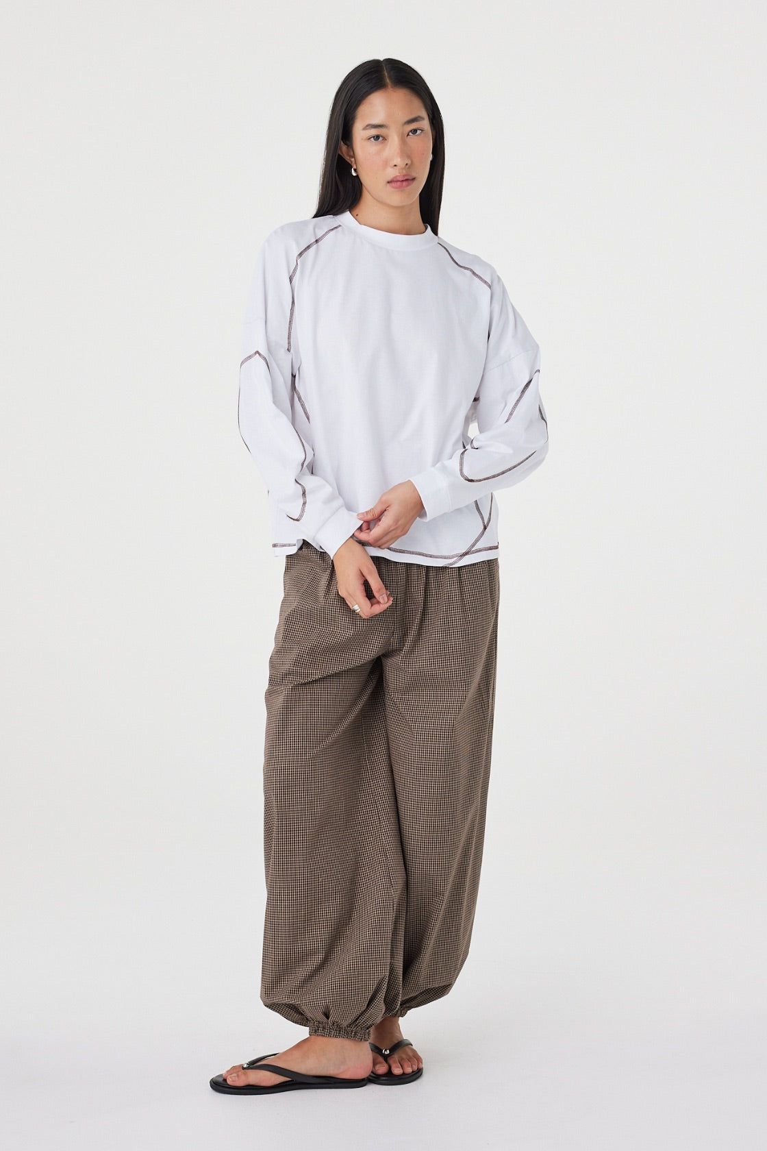 Model wearing the cream long-sleeve tee with brown check trousers and black sandals, showing the dropped shoulders, ribbed cuffs, curved sleeve seams and relaxed drape in a styled front view.