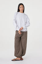 Model wearing the cream long-sleeve tee with brown check trousers and black sandals, showing the dropped shoulders, ribbed cuffs, curved sleeve seams and relaxed drape in a styled front view.