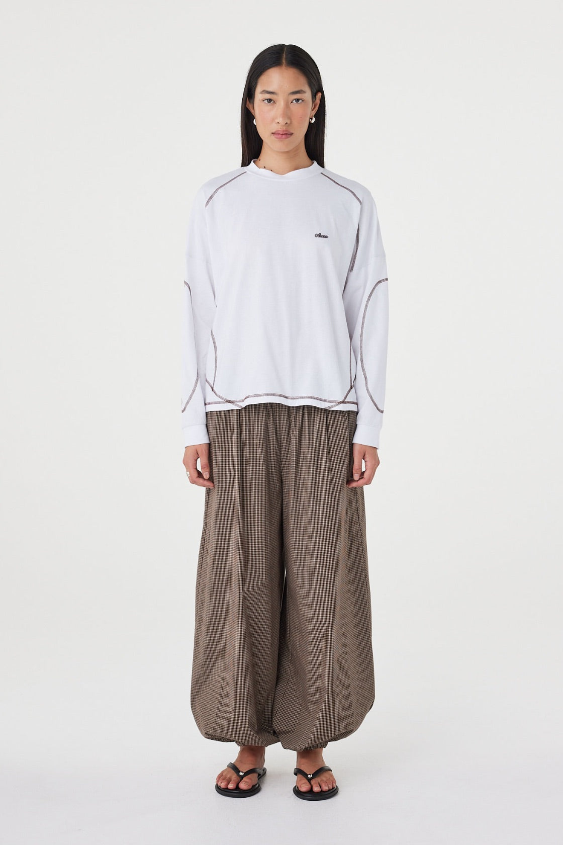 Model wearing the cream long-sleeve tee with brown check trousers, showing the crew neckline, long sleeves, espresso contrast stitching and relaxed off-body silhouette in a full-length front view.