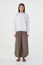 Model wearing the cream long-sleeve tee with brown check trousers, showing the crew neckline, long sleeves, espresso contrast stitching and relaxed off-body silhouette in a full-length front view.