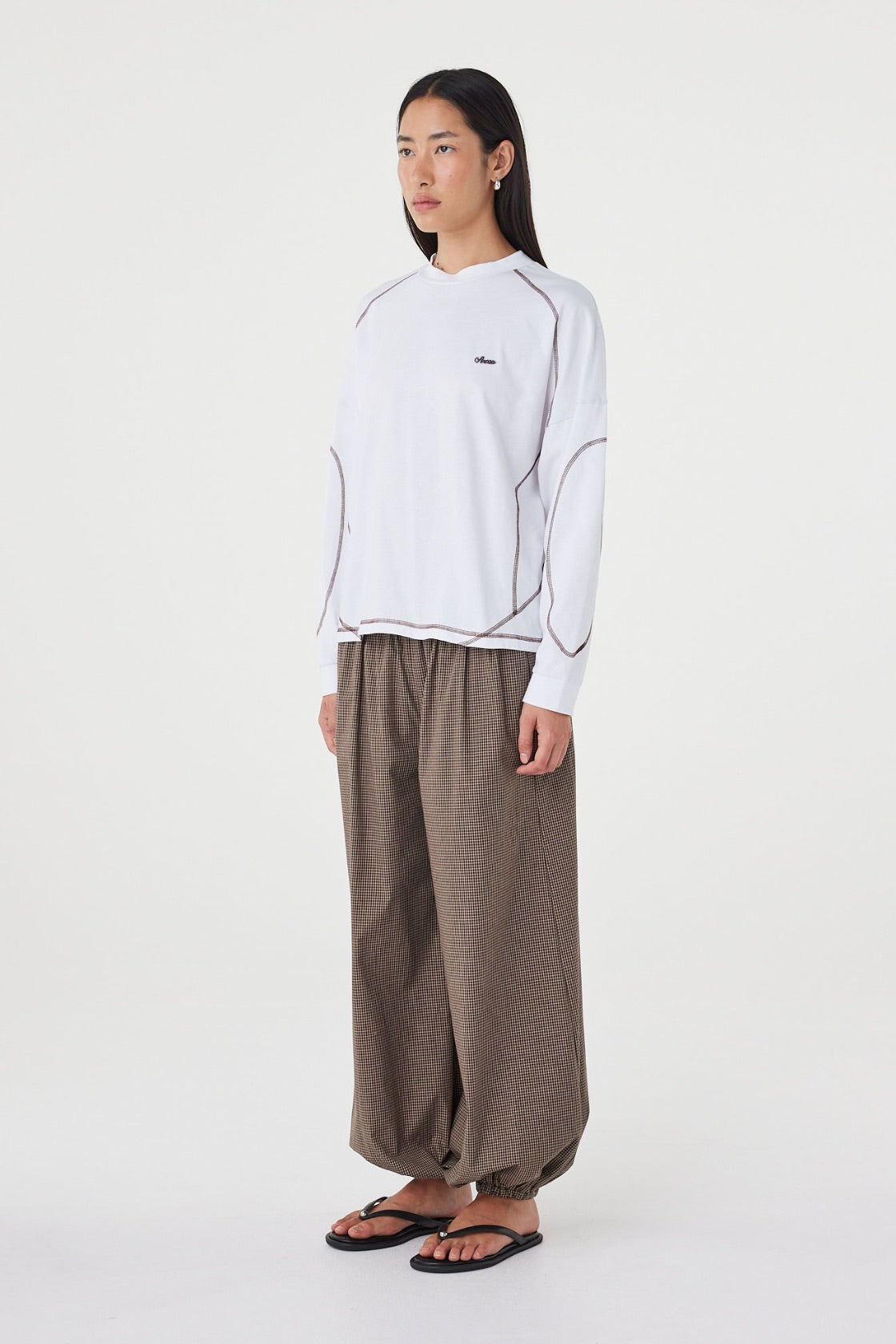 Model wearing the cream long-sleeve tee with brown check trousers and black sandals, showing the crew neckline, dropped shoulders, curved contrast seams and loose hip-length fit in an angled side view.