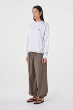 Model wearing the cream long-sleeve tee with brown check trousers and black sandals, showing the crew neckline, dropped shoulders, curved contrast seams and loose hip-length fit in an angled side view.