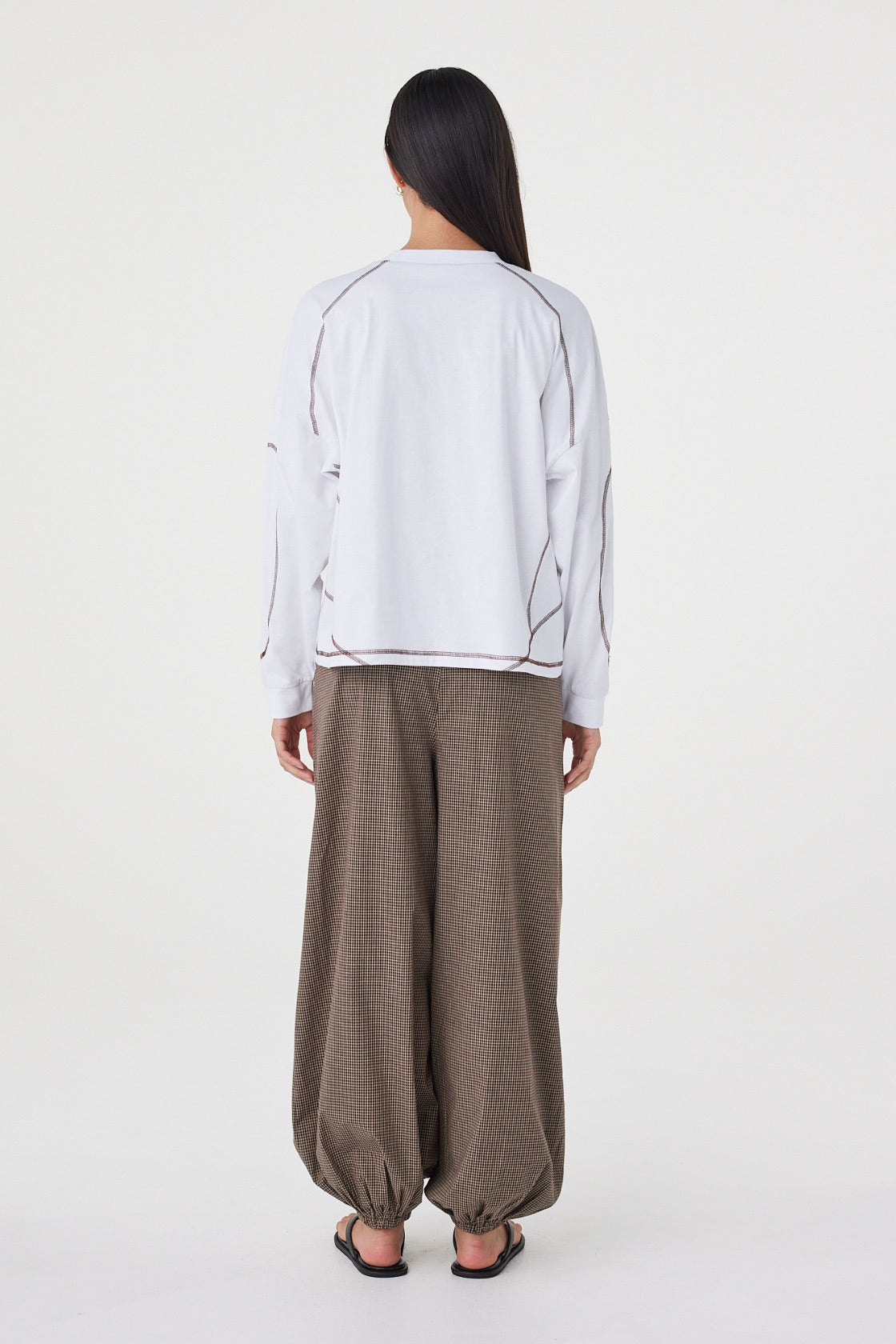 Model wearing the cream long-sleeve tee with brown check trousers and black sandals, showing the dropped shoulders, contrast espresso overlock seams, straight hem and relaxed silhouette in a full-length back view.