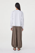 Model wearing the cream long-sleeve tee with brown check trousers and black sandals, showing the dropped shoulders, contrast espresso overlock seams, straight hem and relaxed silhouette in a full-length back view.