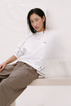 Model wearing the cream long-sleeve tee with brown check trousers, showing the crew neckline, relaxed fit, espresso contrast seams and rolled sleeves in a seated lifestyle view.