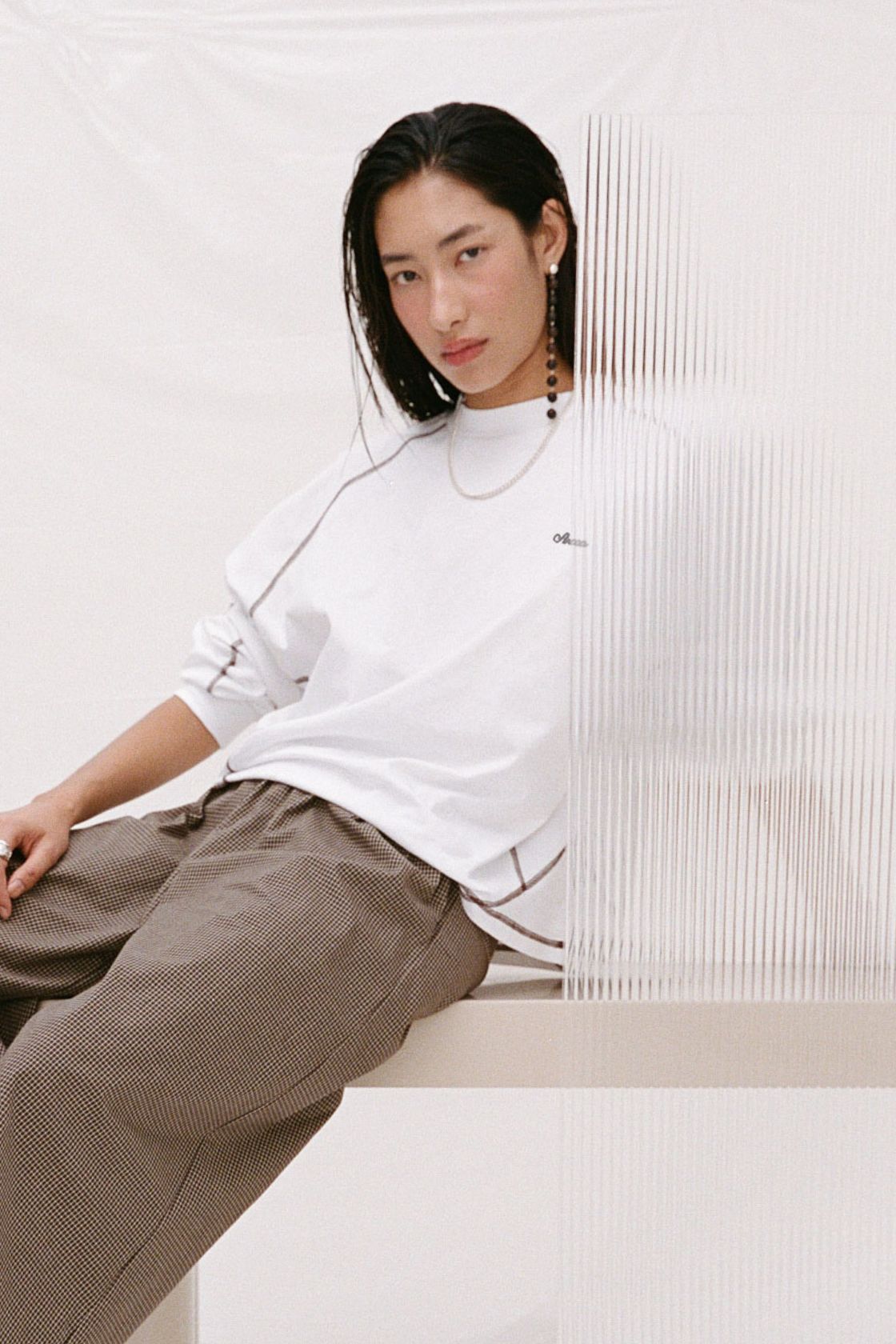 Model wearing the cream long-sleeve tee with brown check trousers, showing the crew neckline, relaxed fit, espresso contrast seams and rolled sleeves in a seated lifestyle view.
