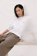 Model wearing the cream long-sleeve tee with brown check trousers, showing the crew neckline, relaxed fit, espresso contrast seams and rolled sleeves in a seated lifestyle view.