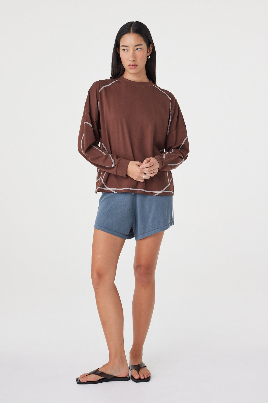 Model wearing the espresso long-sleeve tee with blue shorts and black sandals, showing the relaxed drape, dropped shoulders, curved sleeve seams and contrast stitching in a styled front view.