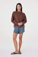 Model wearing the espresso long-sleeve tee with blue shorts and black sandals, showing the relaxed drape, dropped shoulders, curved sleeve seams and contrast stitching in a styled front view.