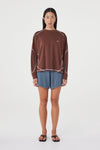 Model wearing the espresso long-sleeve tee with blue shorts, showing the crew neckline, long sleeves, contrast cream overlock stitching and loose off-body silhouette in a full-length front view.