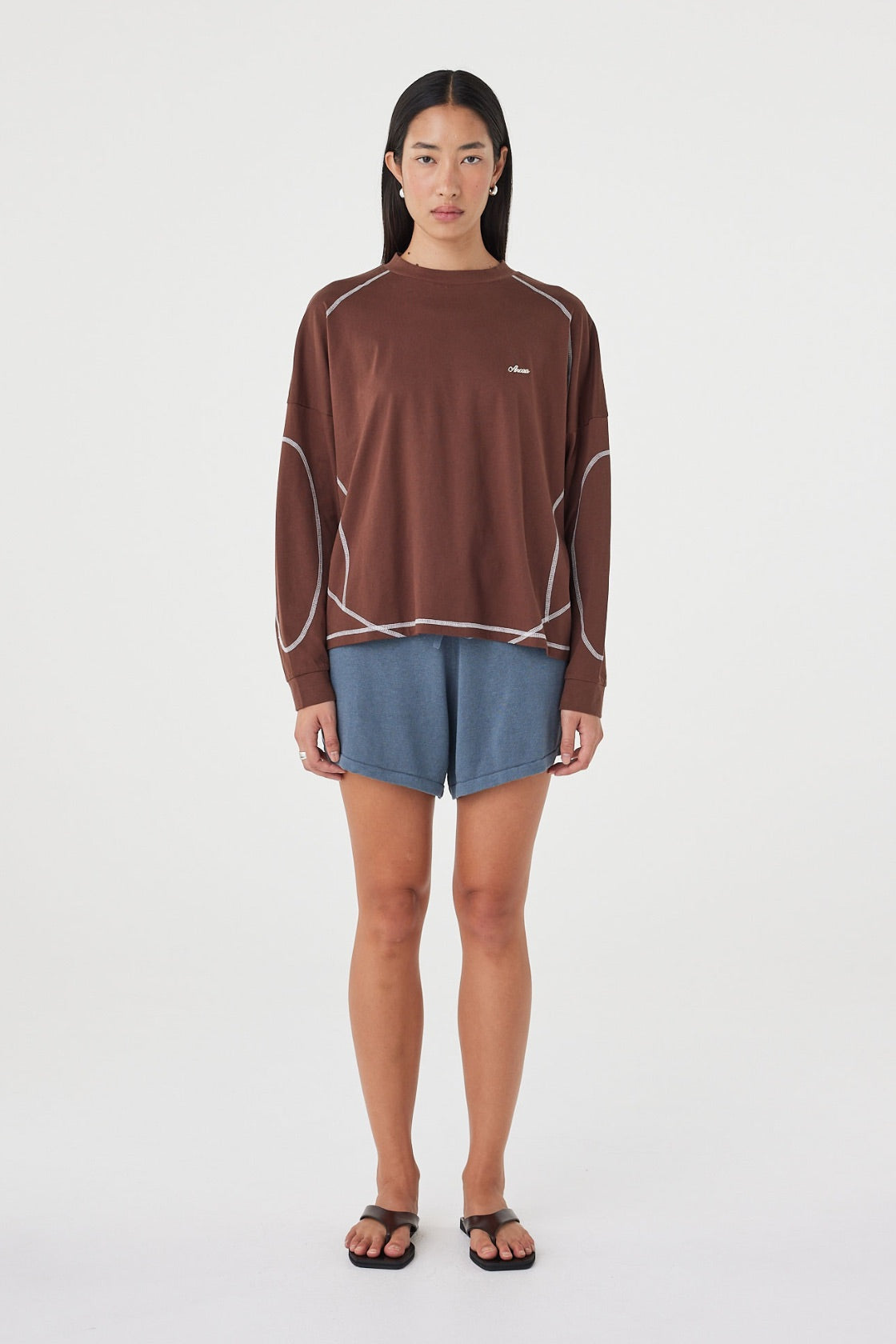 Model wearing the espresso long-sleeve tee with blue shorts, showing the crew neckline, long sleeves, contrast cream overlock stitching and loose off-body silhouette in a full-length front view.