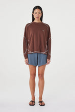 Model wearing the espresso long-sleeve tee with blue shorts, showing the crew neckline, long sleeves, contrast cream overlock stitching and loose off-body silhouette in a full-length front view.