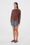 Model wearing the espresso long-sleeve tee with blue shorts and black sandals, showing the crew neckline, dropped shoulders, curved contrast seam detailing and relaxed hip-length fit in an angled front view.