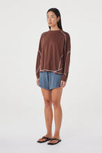 Model wearing the espresso long-sleeve tee with blue shorts and black sandals, showing the crew neckline, dropped shoulders, curved contrast seam detailing and relaxed hip-length fit in an angled front view.