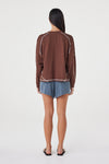 Model wearing the espresso long-sleeve tee with blue shorts and black sandals, showing the dropped shoulders, contrast cream overlock seams, ribbed cuffs and relaxed silhouette in a full-length back view.