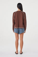 Model wearing the espresso long-sleeve tee with blue shorts and black sandals, showing the dropped shoulders, contrast cream overlock seams, ribbed cuffs and relaxed silhouette in a full-length back view.