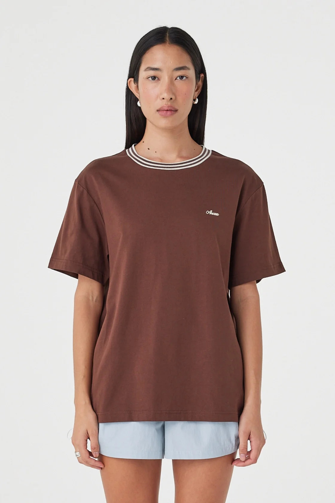 Close-up front view of the espresso oversized tee showing the cream contrast neckline and chest logo, styled with pale blue shorts against a clean white studio background.