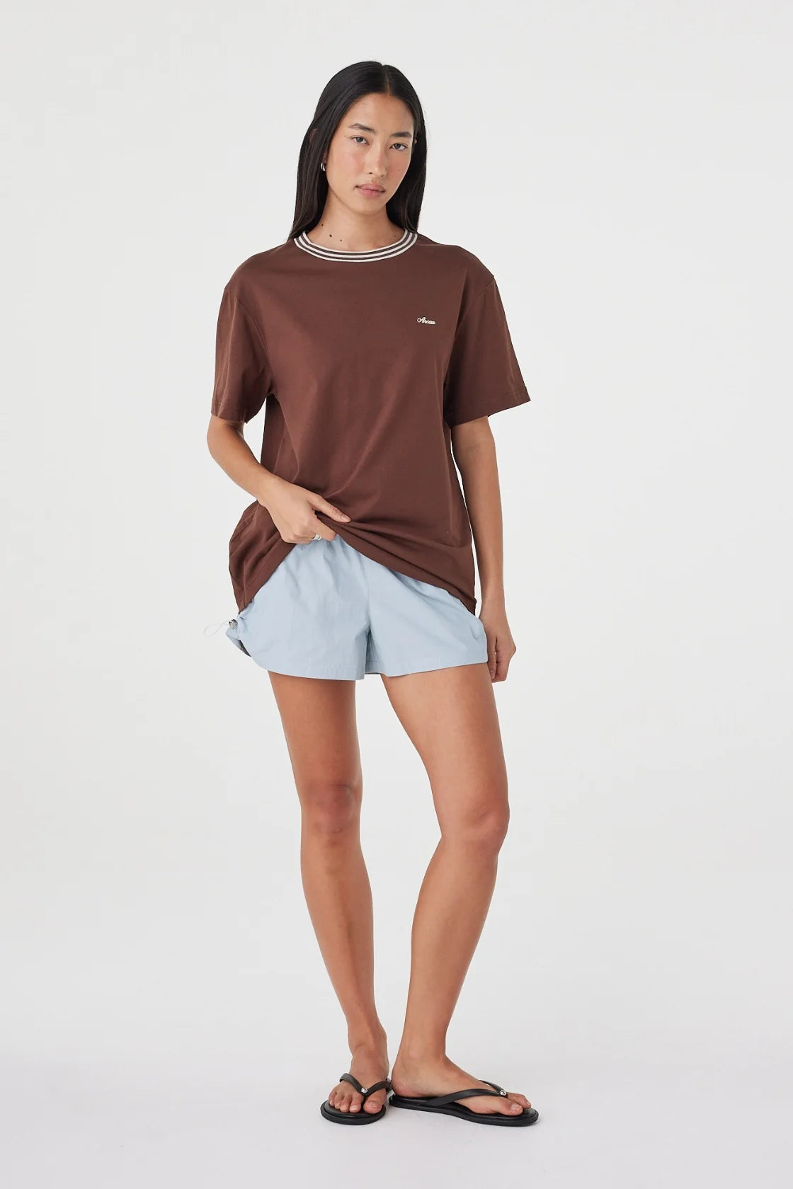 Front angled view of the espresso oversized tee with cream contrast trim, styled with pale blue shorts and black sandals in a relaxed pose against a white studio background.