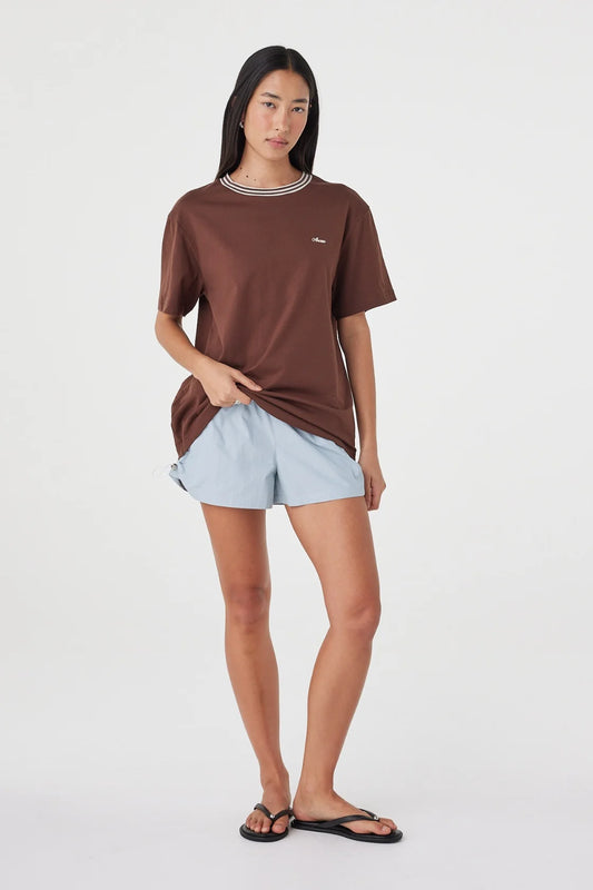 Front angled view of the espresso oversized tee with cream contrast trim, styled with pale blue shorts and black sandals in a relaxed pose against a white studio background.