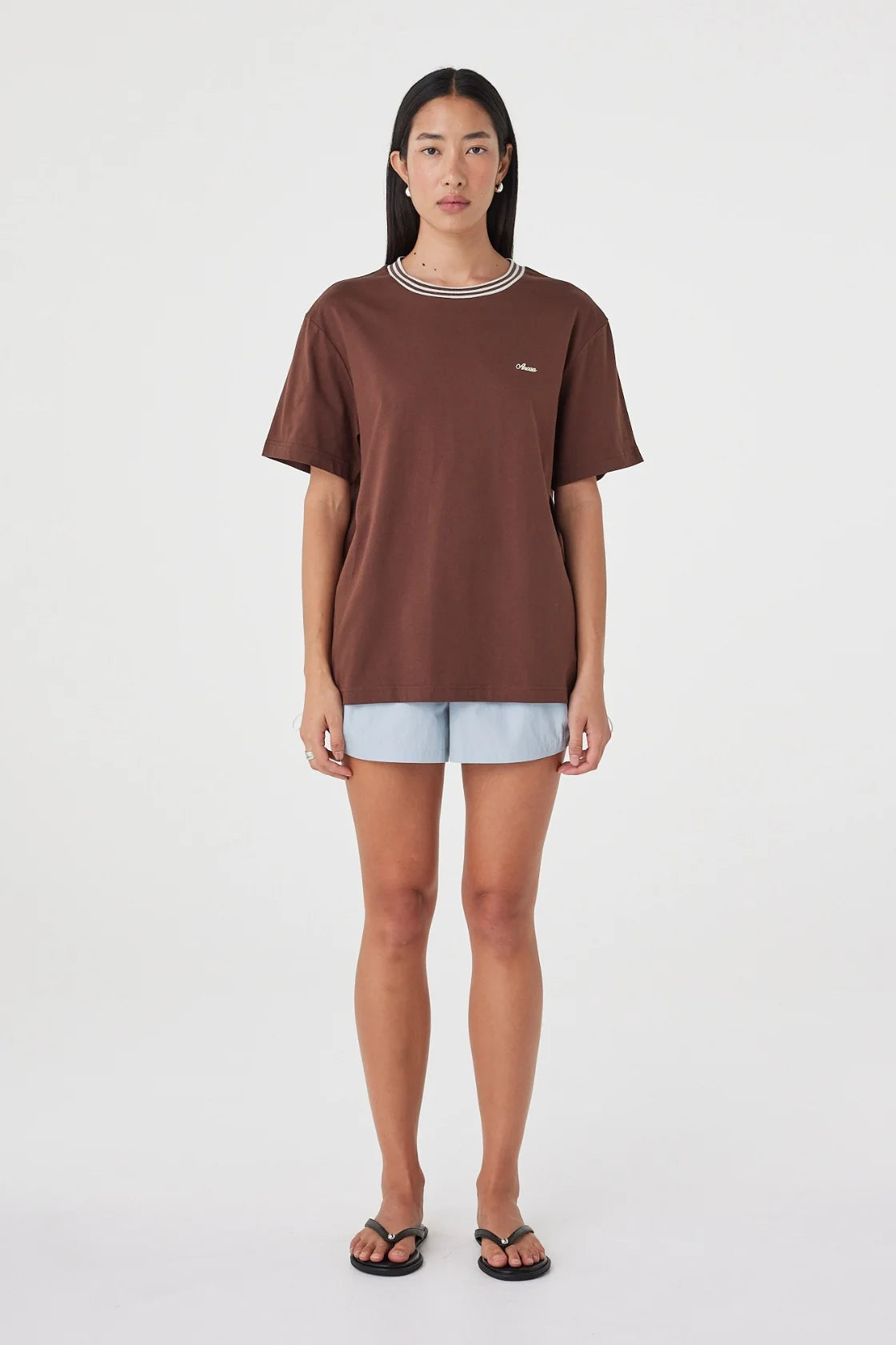 Full-length front view of the espresso oversized tee with cream contrast trim, styled with pale blue shorts and black sandals against a clean white studio background.