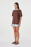 Three-quarter side view of the espresso oversized tee with cream contrast trim, styled with pale blue shorts and black sandals in a minimal white studio.