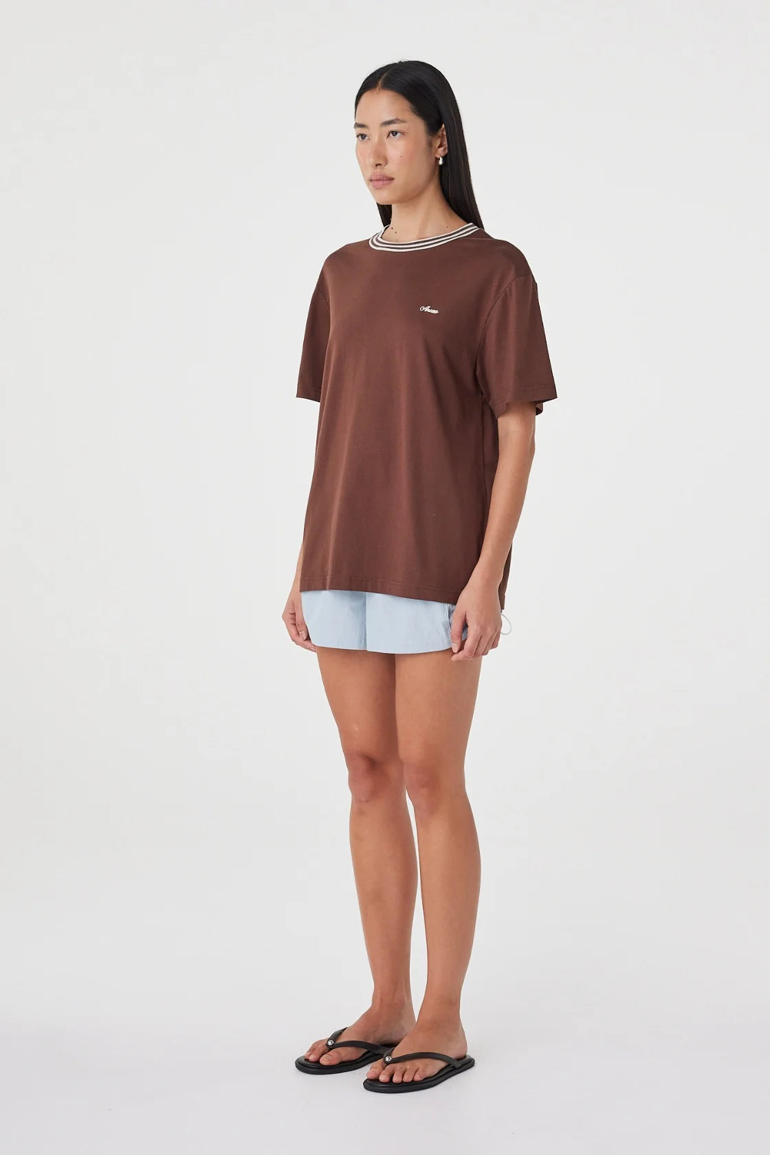 Three-quarter side view of the espresso oversized tee with cream contrast trim, styled with pale blue shorts and black sandals in a minimal white studio.
