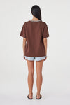 Back view of the espresso oversized tee with cream contrast trim at the neckline, styled with pale blue shorts and black sandals against a clean white studio background.