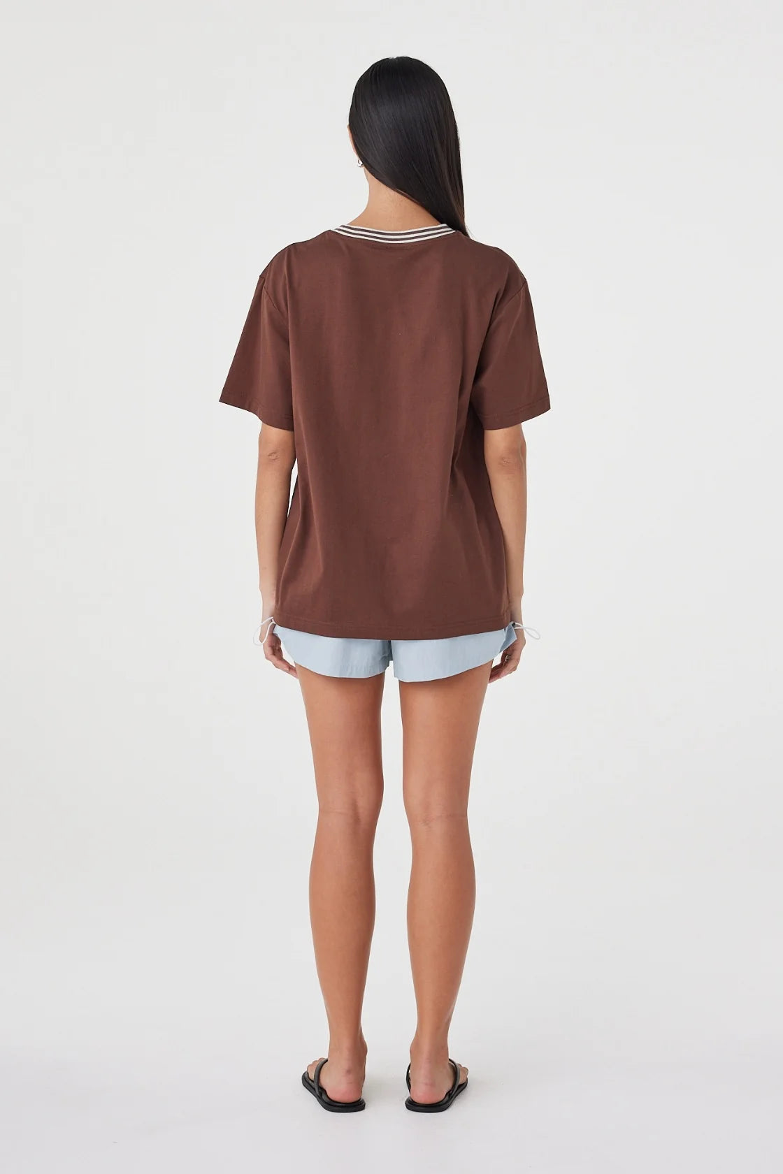 Back view of the espresso oversized tee with cream contrast trim at the neckline, styled with pale blue shorts and black sandals against a clean white studio background.