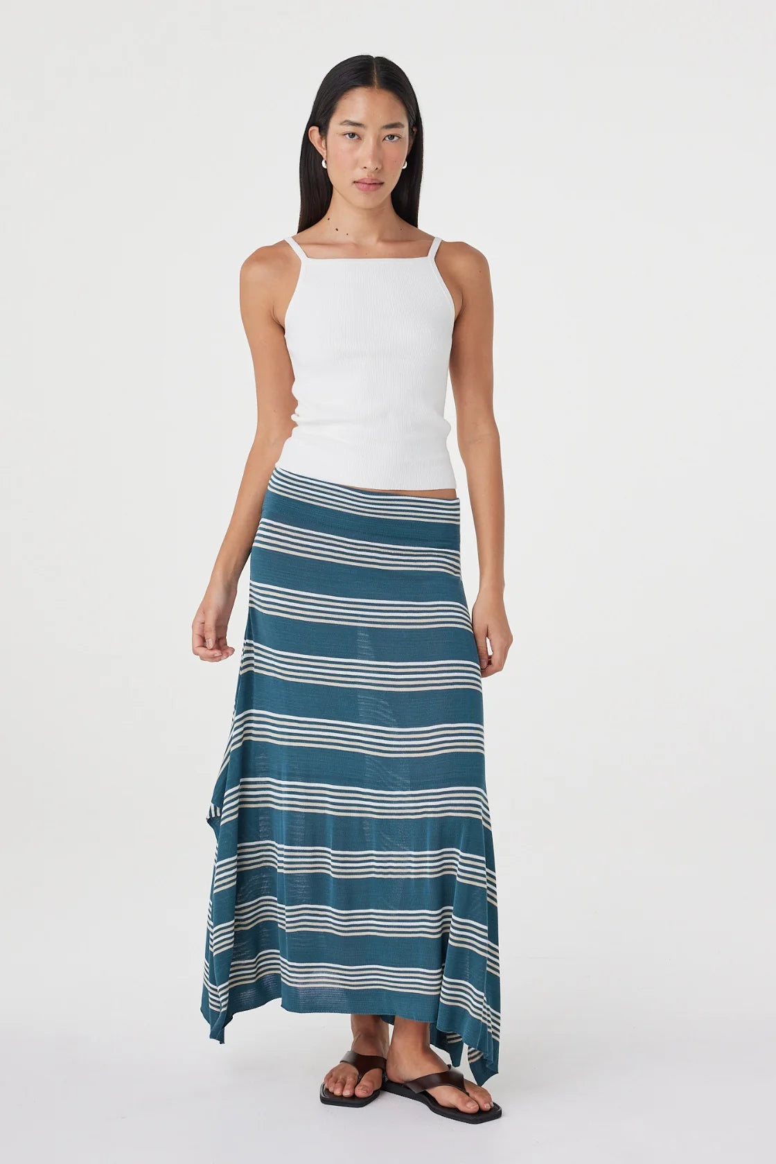 Front angled view of the teal striped maxi skirt showing its asymmetric draped hem, styled with a white sleeveless top and black sandals in a white studio.