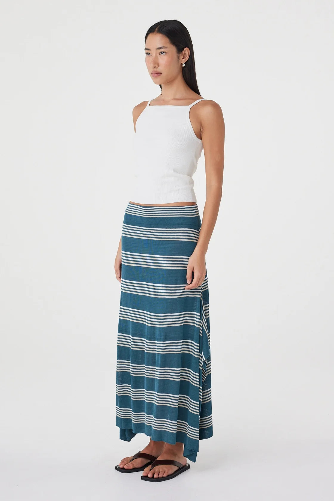 Three-quarter side view of the teal striped maxi skirt styled with a white sleeveless top and black sandals against a minimal white studio background.