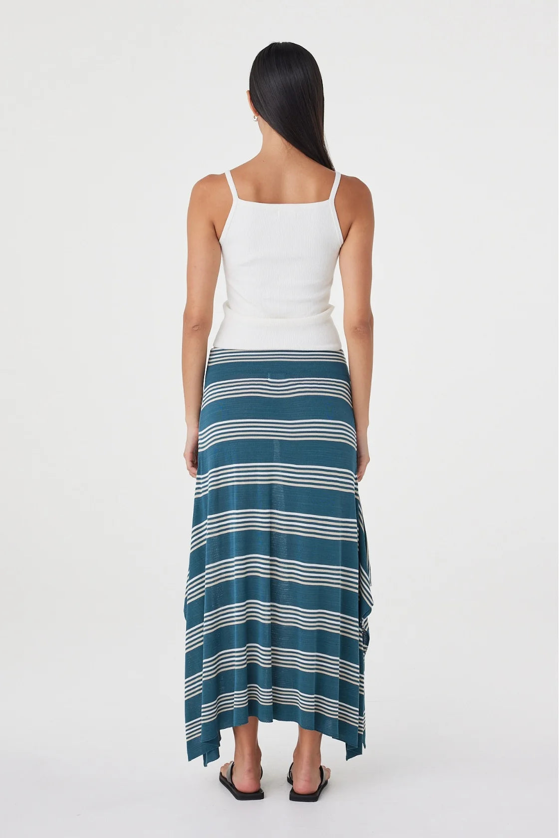 Back view of the teal striped maxi skirt showing the flowing asymmetric hem, styled with a white sleeveless top and black sandals in a clean white studio.