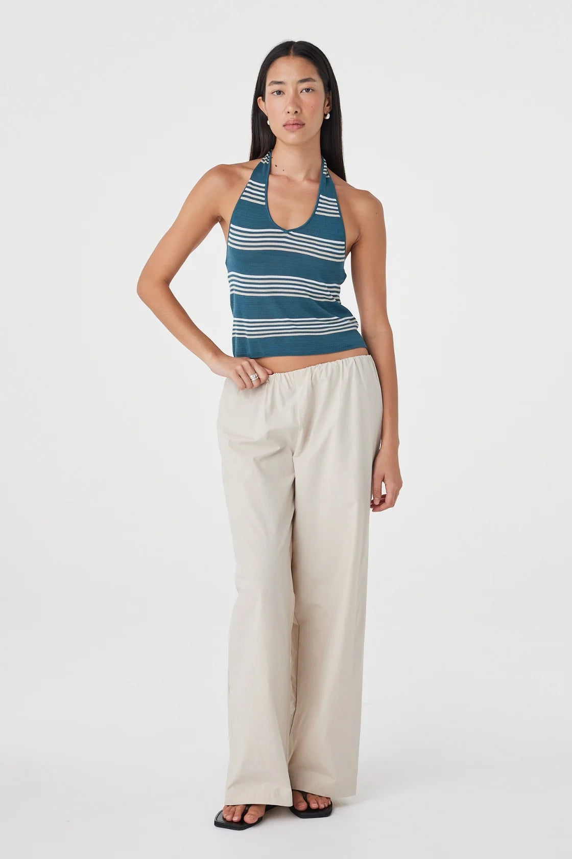 Front angled view of the teal striped halter top styled with cream wide-leg pants and black sandals in a white studio.