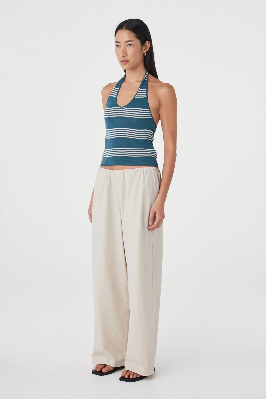 Three-quarter side view of the teal striped halter top styled with cream wide-leg pants and black sandals in a minimal white studio.