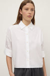 A woman wearing a white cropped button-down shirt with a pointed collar and rolled  long sleeves, shown from the front.