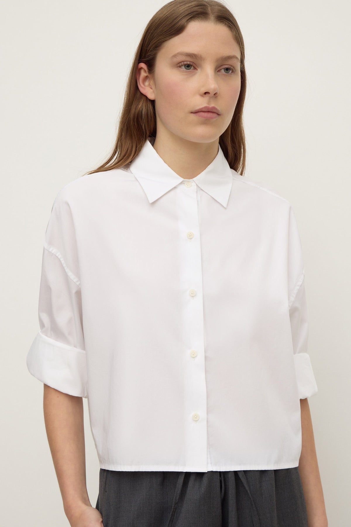 A woman wearing a white cropped button-down shirt with a pointed collar and rolled  long sleeves, shown from the front.