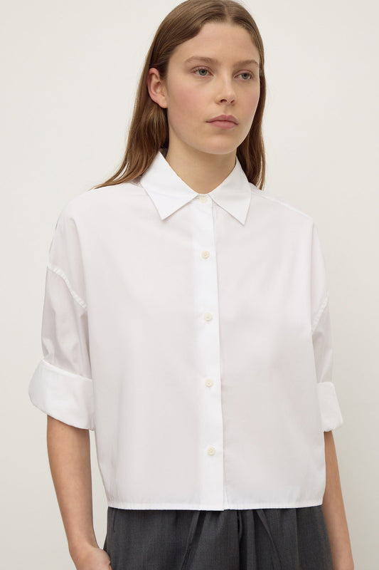 A woman wearing a white cropped button-down shirt with a pointed collar and rolled  long sleeves, shown from the front.