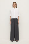 A woman wearing a white cropped button-down shirt with the sleeves rolled to the elbows, shown with wide-leg trousers.