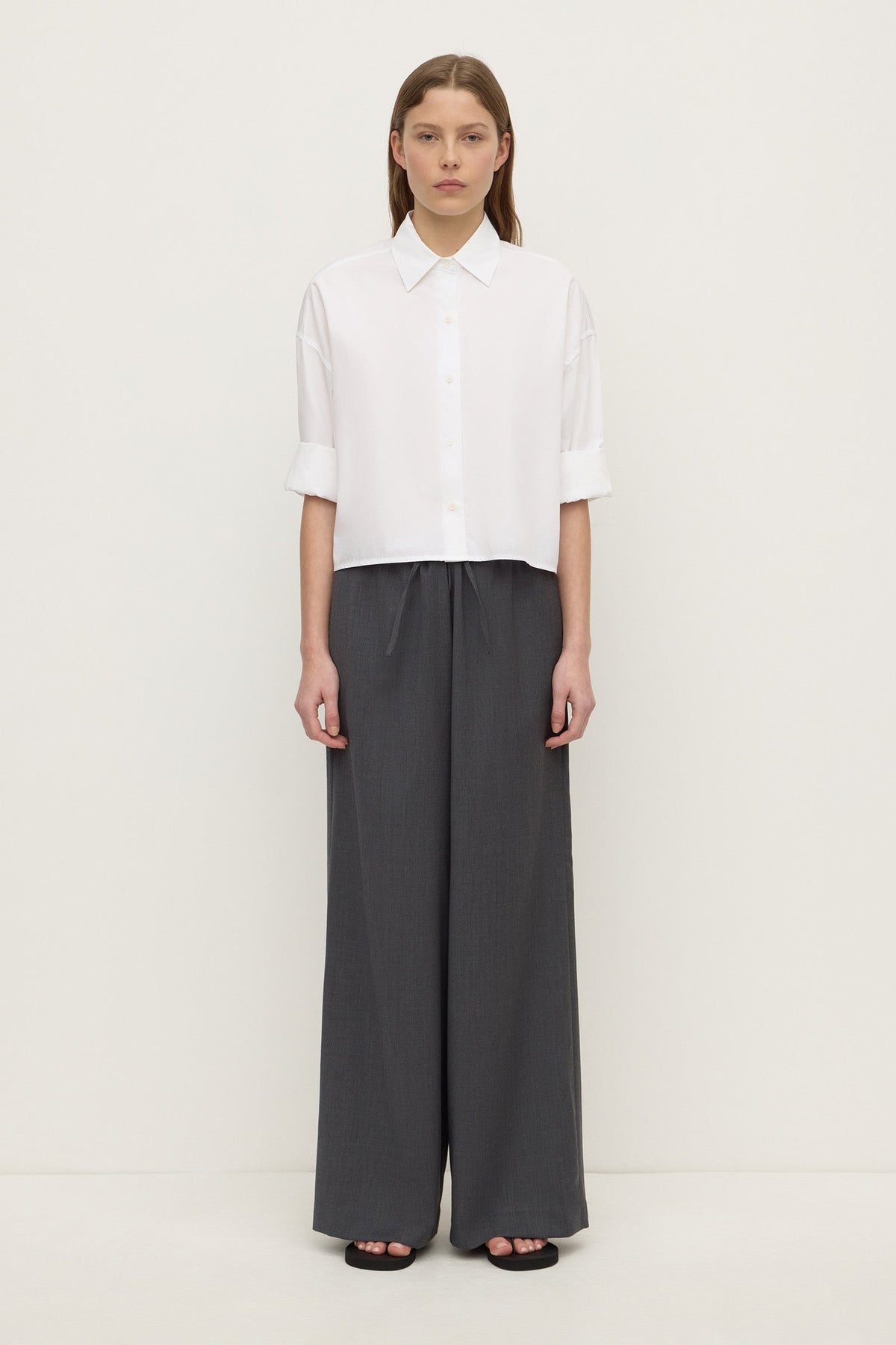 A woman wearing a white cropped button-down shirt with the sleeves rolled to the elbows, shown with wide-leg trousers.