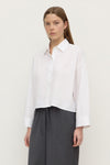 Three-quarter view showing the white shirt's cropped hem, button front and long sleeves.