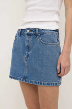 Close-up of the dark stone denim mini skirt with a white fitted tank, showing the mid-rise waistband, front pockets, zip fly and straight mini-length hem
