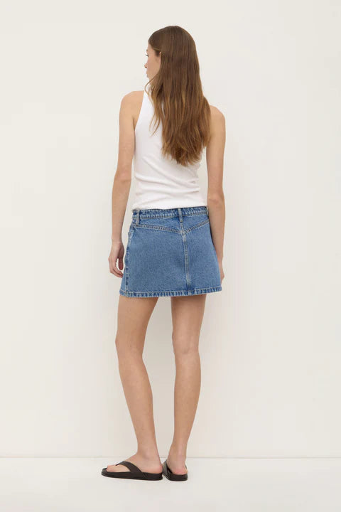 Model wearing the dark stone denim mini skirt with a white tank and black sandals, showing the back yoke, fitted waist and straight mini silhouette, back angled view
