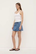 Model wearing the dark stone denim mini skirt with a white fitted tank and black sandals, showing the mid-rise waist, front pockets and clean mini-length shape, angled front view
