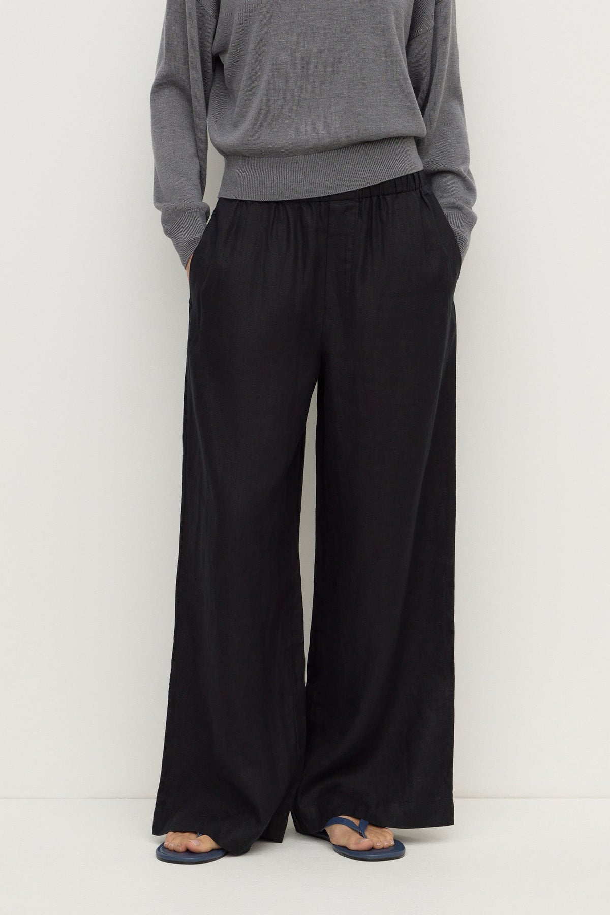 Close-up front view of black wide-leg pants showing the gathered elasticated waistband, side pockets and flowing leg shape.