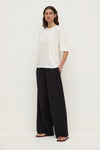 Three-quarter view of a woman wearing an antique white boxy tee with short sleeves and a ribbed neckline, paired with wide-leg black trousers.