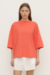 A model stands facing the camera wearing a coral oversized T-shirt with a round ribbed neckline, dropped shoulders and wide elbow-length sleeves, styled with white shorts and black flats.