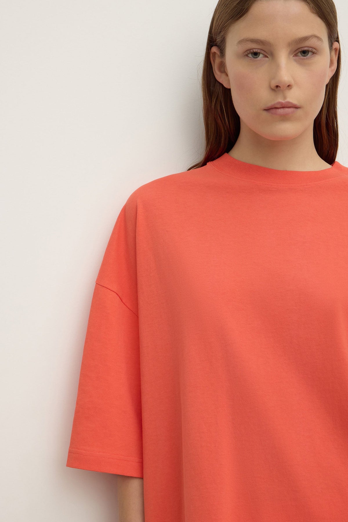 Close-up of a coral T-shirt showing the round ribbed crew neckline, wide shoulder area and loose elbow-length sleeve with visible stitching along the sleeve seam.