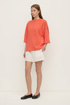 A model stands at a three-quarter angle wearing a coral oversized T-shirt with a loose body, dropped shoulders and wide elbow-length sleeves, paired with white shorts and black flats.