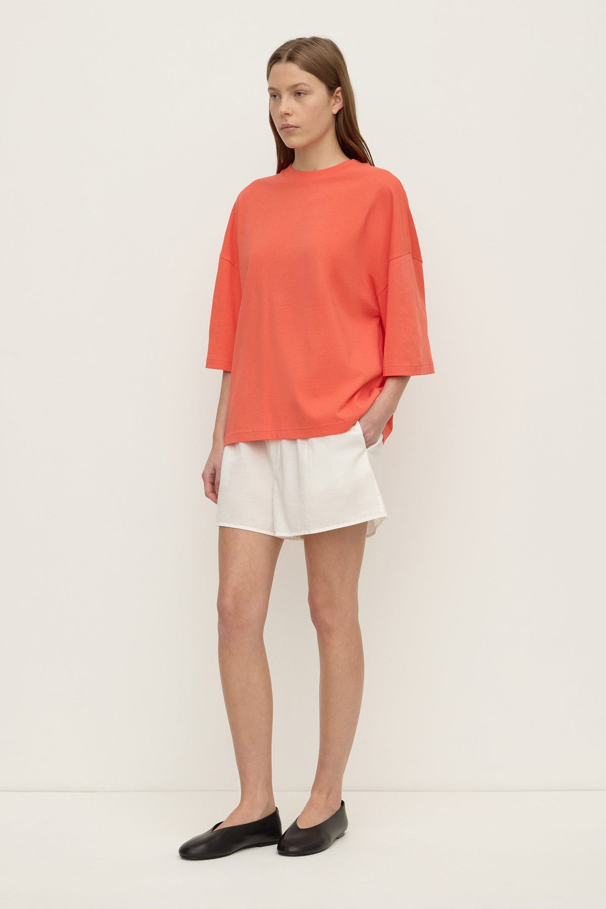 A model stands at a three-quarter angle wearing a coral oversized T-shirt with a loose body, dropped shoulders and wide elbow-length sleeves, paired with white shorts and black flats.
