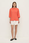 A model stands facing forward in a coral oversized T-shirt with a round neckline, relaxed body, dropped shoulders and wide elbow-length sleeves, paired with white shorts.